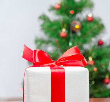 White gift box with red ribbon on background of Christmas tree