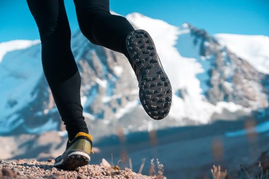 Athlete runner legs run on a trail along a rocky path close-up. Fitness and warm-up, young man with snow-covered mountain peaks in the background and open space around him at sunset.