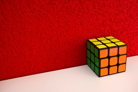 Solved puzzle speed cube on white wooden shelf near red colored wall, colorful rubiks cube with copy space mind challanging retro design