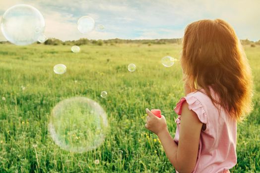 Little girl in pink dress blowing soap bubbles on summer meadow at sunny day, rear view. Image with sunlight effect