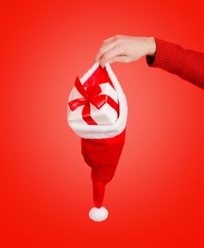 Human hand is holding santa hat with gift box on a red background