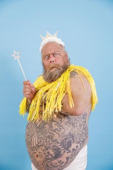 Doubting bearded plus size man with toy crown and magic stick wearing decorative cape poses on light blue background in studio