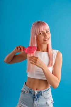 Smiling woman brushing her smooth natural long pink hair with hair comb while standing isolated over blue studio background. Beauty, hair care concept