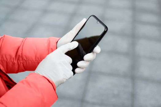 Young woman holding a mobile phone in her hands wearing latex protective gloves at the street in public place. Coronavirus spreading precautions measures concept.