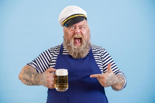 Bearded man with overweight wearing sailor suit with apron points onto mug of tasty beer by finger on light blue background in studio