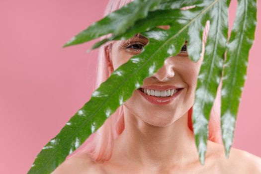 Portrait of happy woman with pink hair smiling at camera, hiding behind green plant leaf, posing isolated over pink studio background. Beauty, skin care, natural cosmetics concept