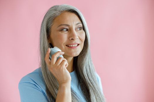 Attractive silver haired Asian woman uses sonic facial cleansing brush posing on pink background in studio. Mature beauty lifestyle