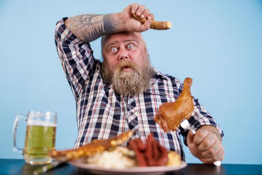 Tired middle aged obese man mops brows suffering from overeating at table with fat food and beer on light blue background in studio