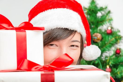 Young woman in santa hat looks out from behind presents on a background of Christmas tree