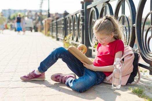 Child girl student wearing glasses with backpack is reading book. Urban background, river, sky