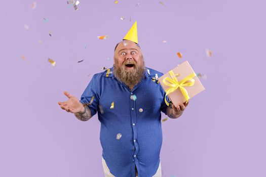 Positive emotional plus size man in tight blue shirt and party hat holds gift box standing under confetti shower on purple background in studio