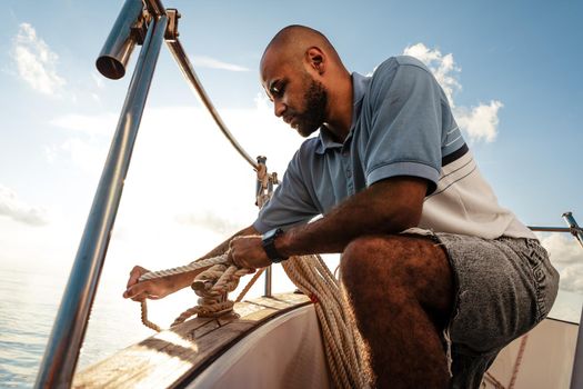 Young african american sailor tying ropes on sailboat in the sea on sunset, close up