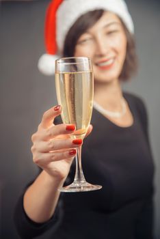 Happy woman in santa hat is giving tumbler with champagne on a gray background