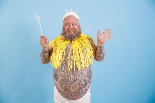 Joyful mature bearded plump man with toy crown, magic stick and yellow cape stands on light blue background in studio