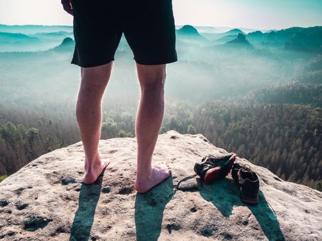 Barefoot slender legs with hairy calves of a male runner standing next to removed sweaty running shoes on a rocky edge above a long deep vally in nature park.
