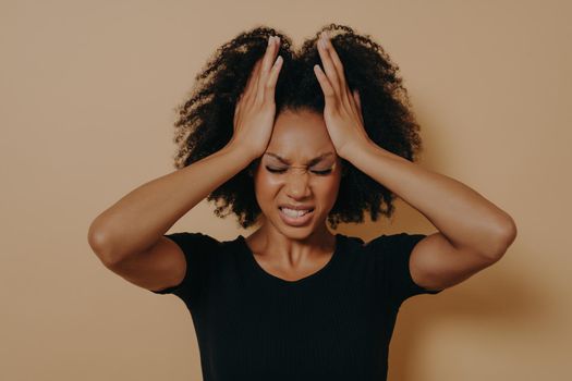 Shocked panic African American girl dressed in black T-shirt holding hands on head and screaming in despair and frustration, with eyes closed and teeth clenched with anger, posing in studio background