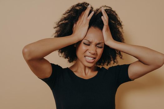 Shocked African American stressed girl holding hands on head and showing despair and frustration while having terrible headach, her eyes closed in pain and teeth clenched. Negative women emotions