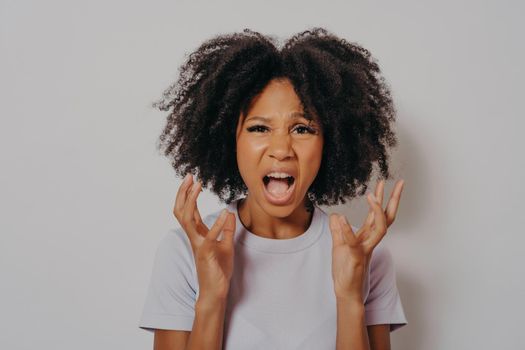 Young beautiful African American woman with curly hair wearing casual tshirt, standing angry and mad, raising hands up frustrated and furious while shouting with anger. Rage and aggressive concept