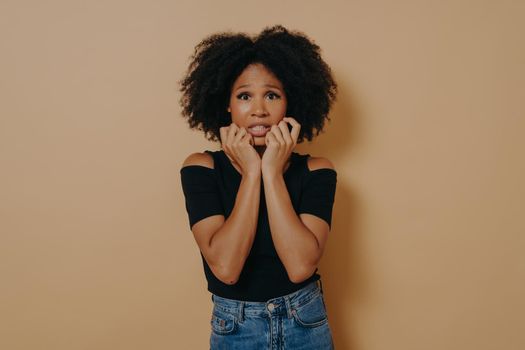 Woman about to panic hearing shocking worried news. Portrait of intense nervous and anxious dark-skinned female clenching fists opening mouth looking concerned over dark beige wall with copy space