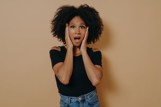 Photo of shocked pretty dark skinned curly haired lady holding hands on cheekbones not believe becouse of discount low prices, wear trendy black tshirt isolated over dark beige studio background