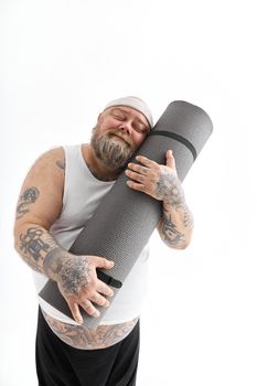 Happy fat man with big belly and tattos in sports wear is huging exercise mat and smiles