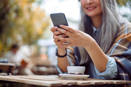 Happy grey haired mature lady writes message on phone sitting at table with cup on outdoors cafe terrace on autumn day closeup