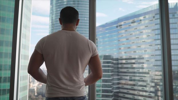 Athletic man looking out the window in the morning. Handsome young guy enjoying beautiful view on a sunny day. Rear back view Close up movement to the window. Athlete on the background of window windows.