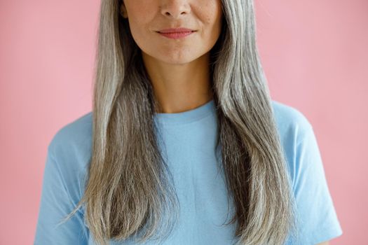 Positive middle aged woman with long hoary hair in blue t-shirt on pink background in studio closeup. Mature beauty lifestyle