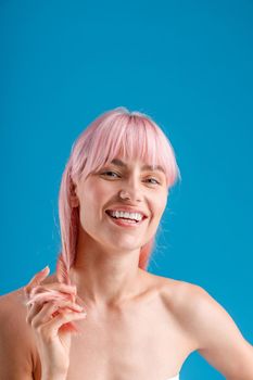 Portrait of beautiful naked woman smiling at camera, touching her pink straight hair while posing isolated over blue studio background. Beauty, skin care concept
