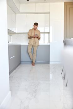 Hadsome smiling man is chilling on the minimalistic kitchen with smartphone