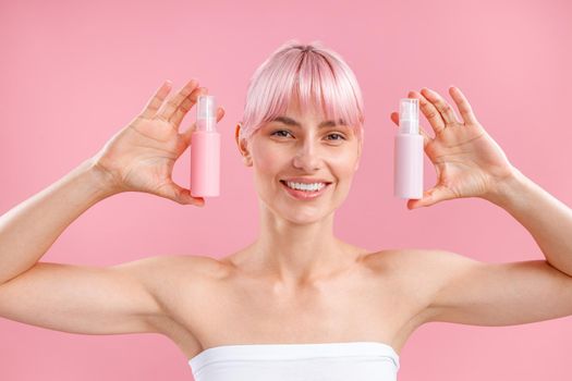 Portrait of excited young woman with pink hair smiling and showing two plastic bottles with beauty products, posing isolated over pink background. Beauty, spa, body care concept