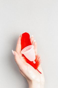 Close up of woman hand holding menstrual cup with red feather over grey background. Periods hygiene product, zero waste alternatives