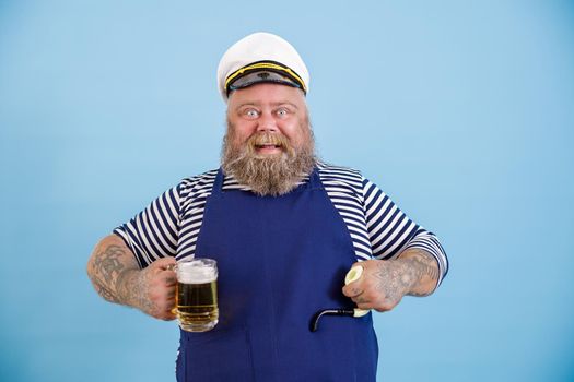 Funny emotional male person with overweight in sailor costume with apron holds smoking pipe and mug of beer on light blue background in studio