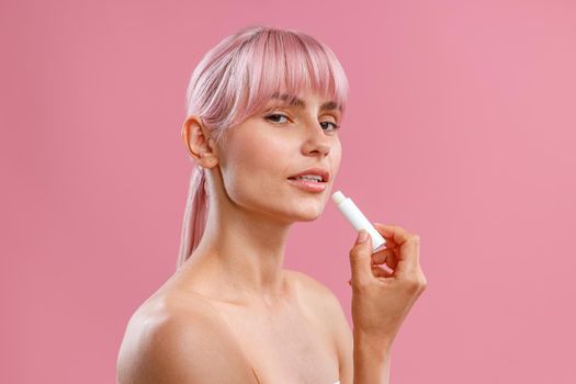 Portrait of beautiful young woman with naked shoulders holding lip balm near her lips isolated over pink background. Beauty, lip care concept