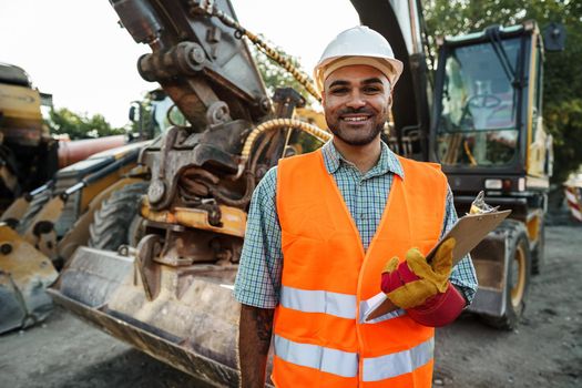 Young man engineer in workwear standing in construction site with clipboard, close up portrait