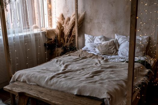 Home interior with four poster bed. There are many white and beige pillows and bedspreads on the bed. A bouquet of dried flowers stands nearby. The bed is decorated with a garland