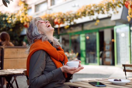 Cheerful senior lady with glasses and cup of tasty coffee laughs throwing back her head at table on outdoors cafe terrace on autumn day