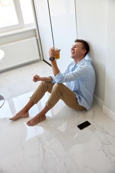 Caucasian hadsome young man in blue shirt is sitting on the floor and chilling at modern kitchen