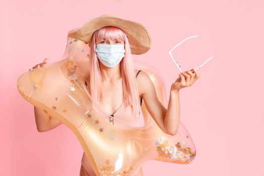 Woman in hat and swimsuit with inflatable rubber ring wearing hygienic face mask to prevent contagious coronavirus on resort beach, posing isolated over pink background. Summer vacation, Covid19