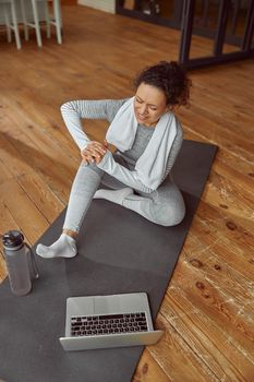 Top view of woman sitting on mat and suffering pain in leg while doing workout with laptop