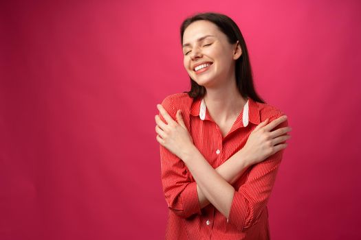 Joyful smiling young woman hugging herself against pink background, close up
