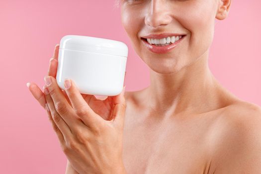 Close up shot of young woman holding white jar with moisturizing body lotion after shower isolated over pink background. Beauty, spa, body care concept