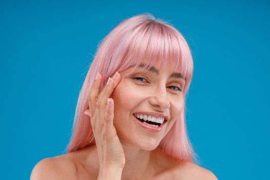 Portrait of smiling young woman with pink hair touching her perfect skin, looking at camera, posing isolated over blue studio background. Beauty, skin care concept