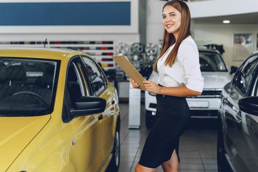 Attractive young female car dealer standing in showroom near a new car