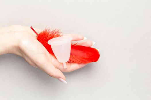 Close up of woman hand holding menstrual cup with red feather over grey background. Periods hygiene product, zero waste alternatives