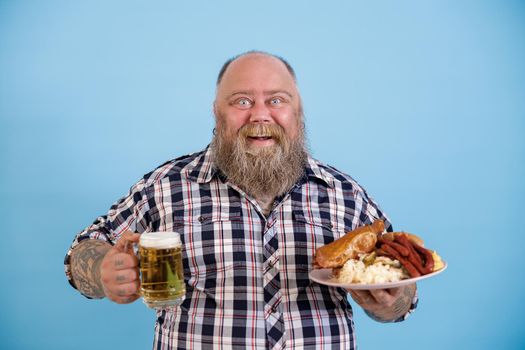 Positive middle aged bearded man with overweight holds plate of greasy food and mug of beer standing on light blue background in studio