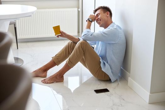 Caucasian hadsome young man in blue shirt is sitting on the floor and chilling at modern kitchen