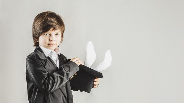 Young magician hiding a toy bunny in his top hat over grey background. Magical performance concept