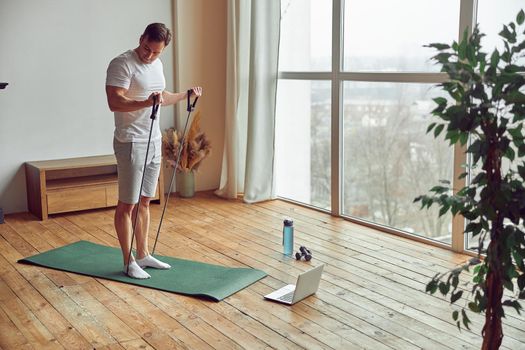 Athletic guy is using resistance bands for biceps workout in front of laptop during quarantine