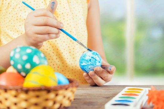 Child is painting egg in blue color for Easter holiday, face is not visible